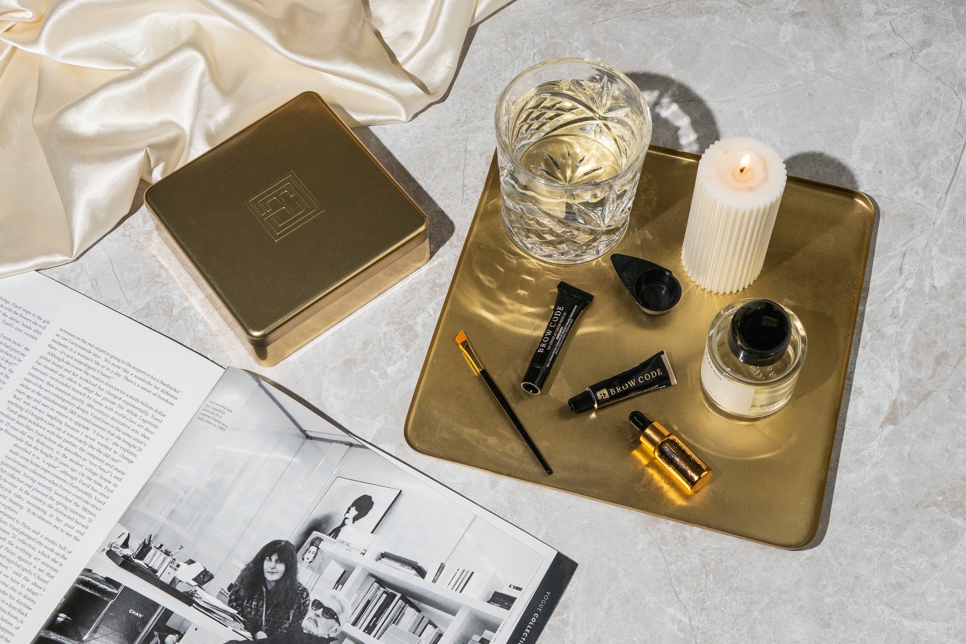 Ein goldenes Tablett enthält ein Getränk, eine Kerze, Make-up, Brow Code AT HOME KIT Augenbrauenfarbe (2 Farben), neben einer aufgeschlagenen Zeitschrift. Blashy Shop