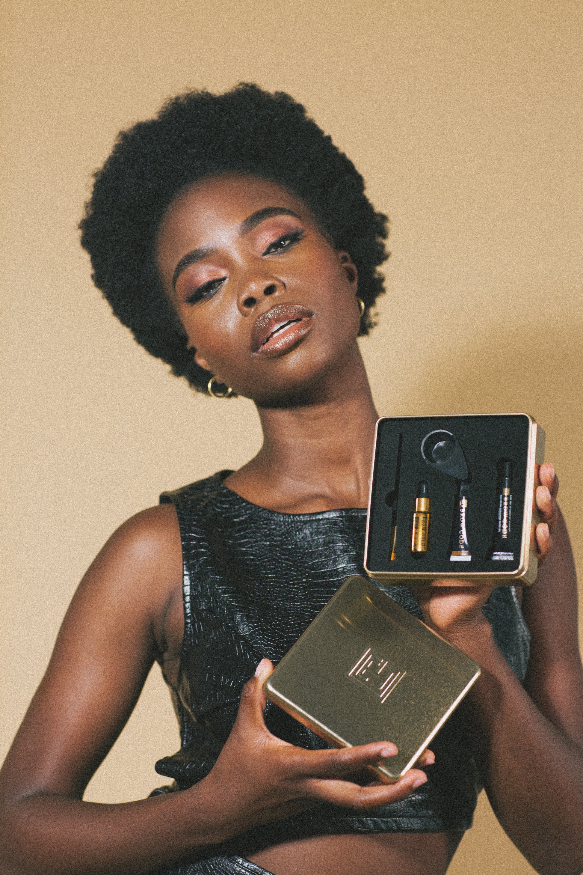 Eine Frau hält eine geöffnete goldene Schachtel mit dem Brow Code AT HOME KIT Augenbrauenfarbe (2 Farben) vor einem beigen Hintergrund. Blashy Shop