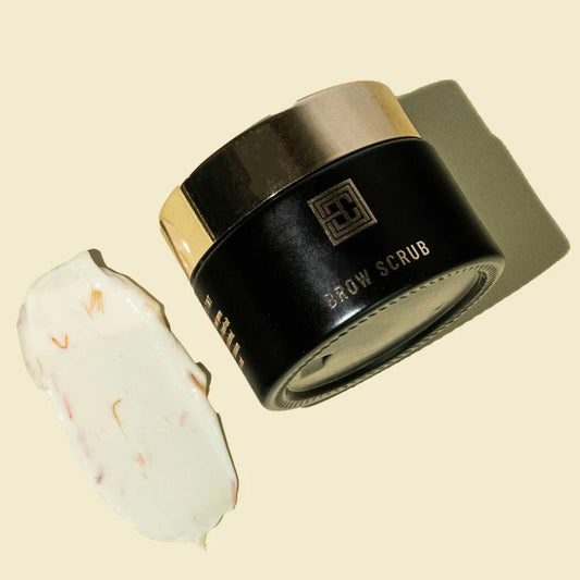 Ein schwarz-goldener Tiegel Brow Code Augenbrauen Peeling 50ml steht neben einem cremigen Abstrich auf beigefarbenem Hintergrund. Blashy Shop