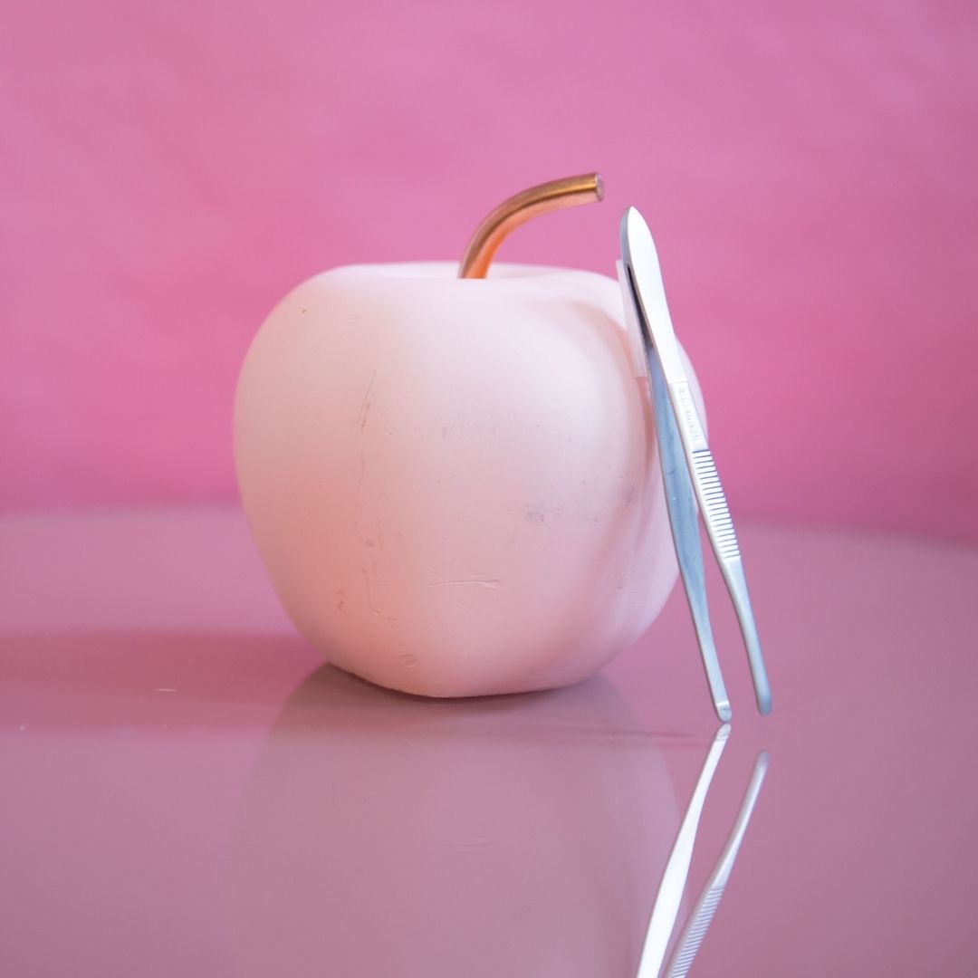 Ein zartrosa Apfel mit metallischem Stiel sitzt neben der Blashy EASY TWEEZY Augenbrauen-Pinzette - rund auf rosa. Blashy Shop