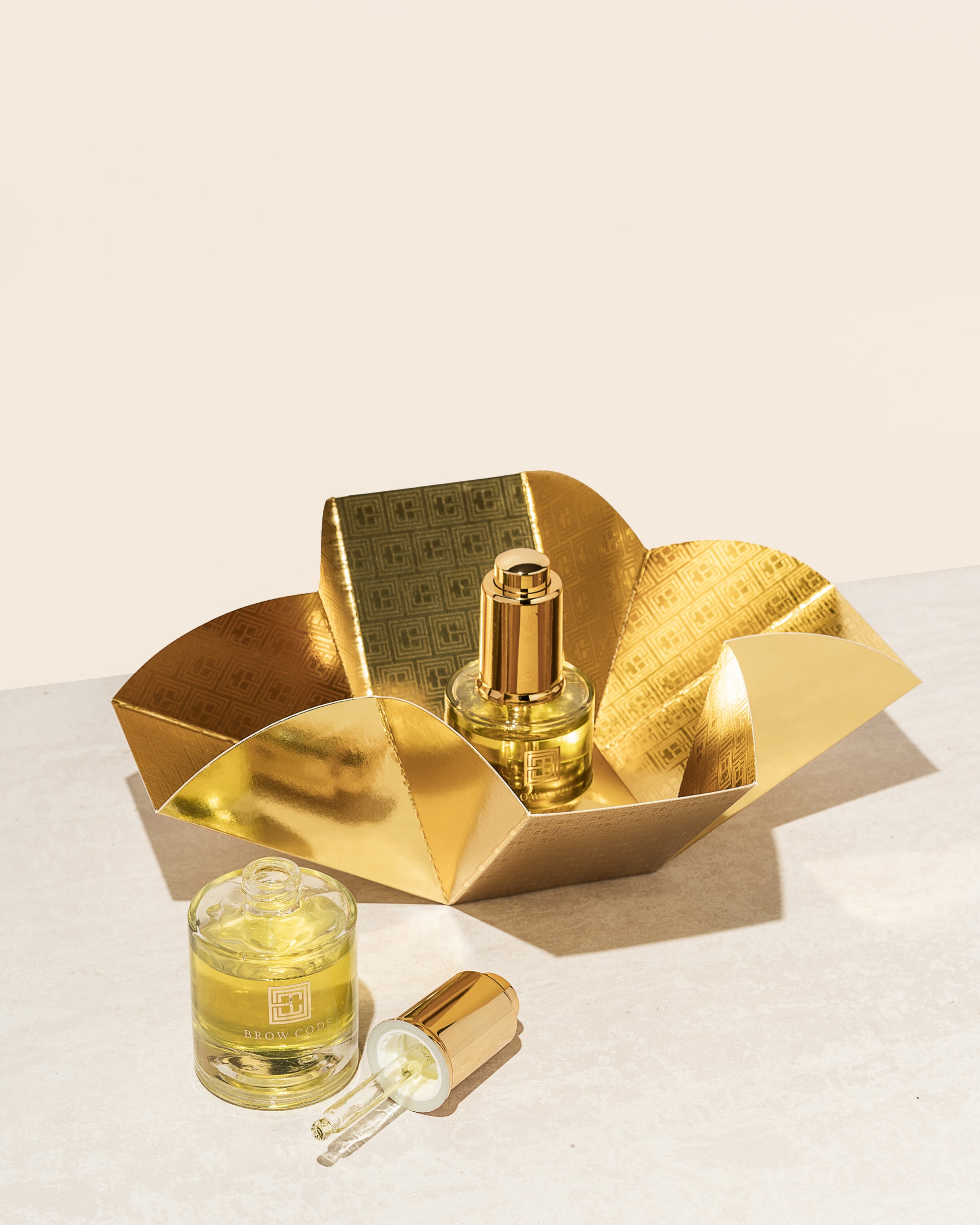 Zwei Flaschen Brow Code Brow Gold Oil pflegendes Wachstumsöl, 30ml, mit Tropfer und Goldhalter auf einer hellen Oberfläche. Blashy Shop