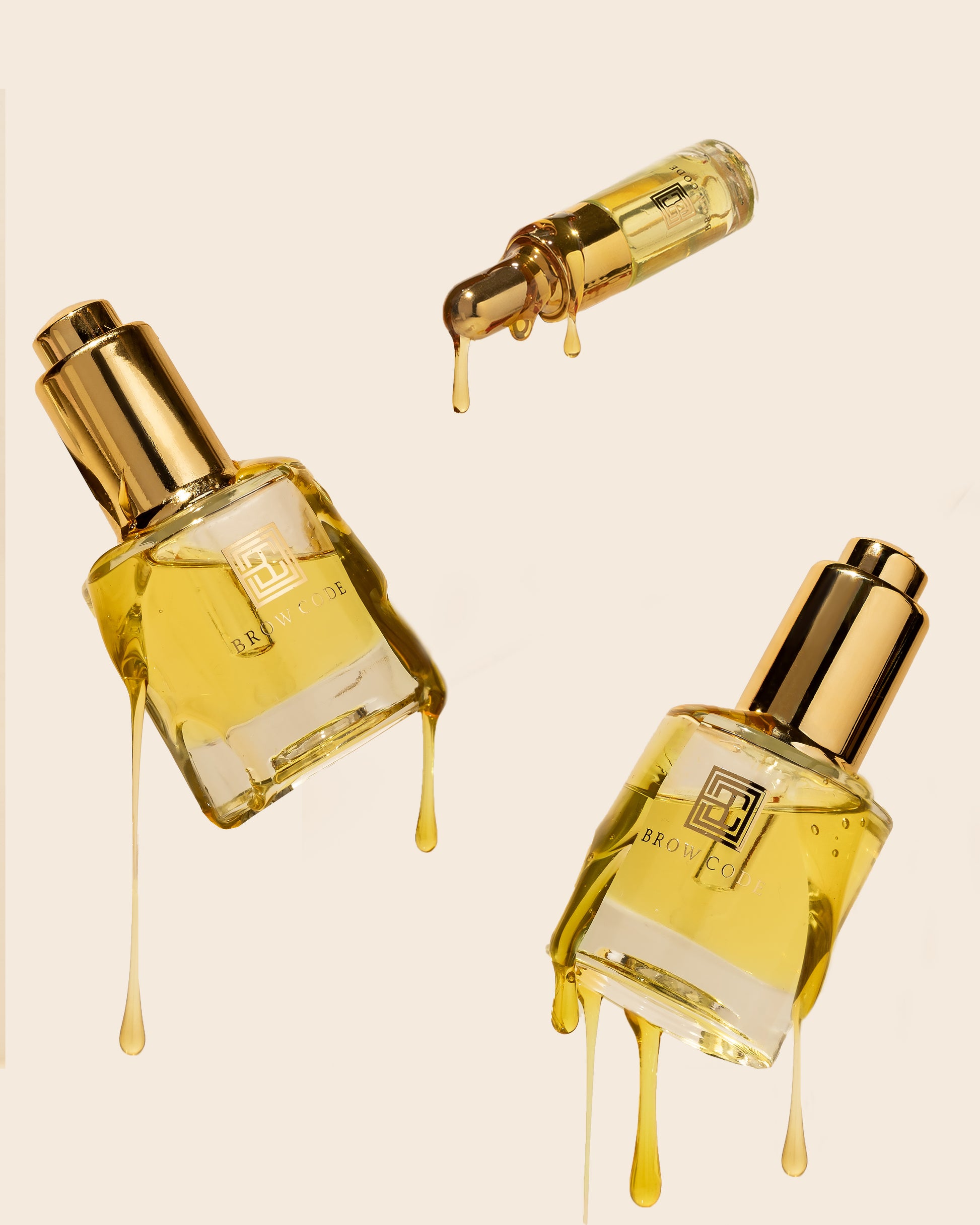 Drei Brow Code Brow Gold Oil pflegendes Wachstumsöl, 30ml Flaschen mit tropfender Flüssigkeit auf einem beigen Hintergrund. Blashy Shop