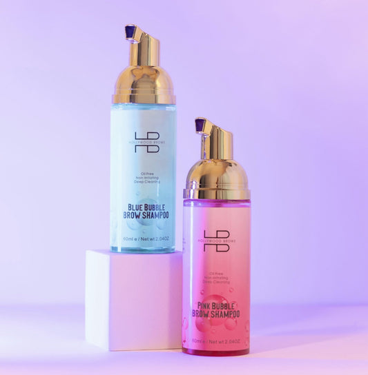 Zwei Flaschen Hollywood Brows Bubble Brow Shampoo Schaum in pink & blau mit goldenen Pumps, auf weißen Blöcken und lila Hintergrund. Blashy Shop