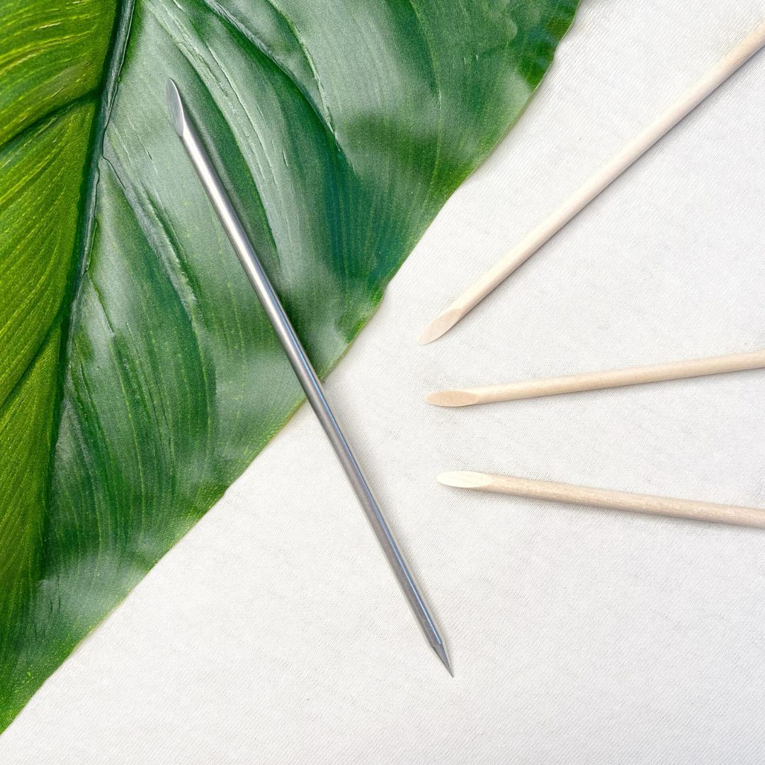 Ein BWR Brow Tool von blashyshop.com und drei hölzerne Nagelhautstäbchen liegen auf einer weißen Fläche neben einem grünen Blatt. Blashy Shop