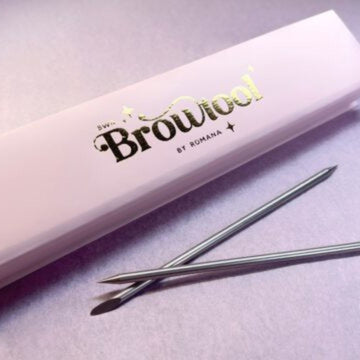 Eine rosa blashyshop.com Brow Tool Box und zwei BWR Edelstahl Wachsspatel ruhen auf einer hellvioletten Fläche. Blashy Shop