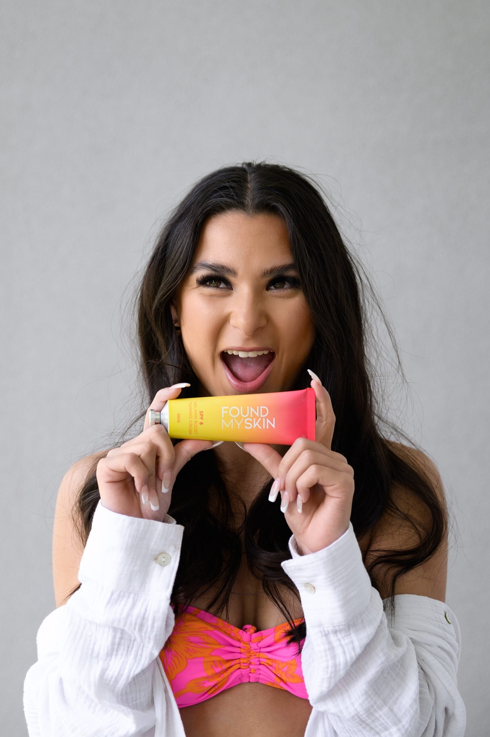 Frau im rosa Bikini-Oberteil lächelt und hält FOUND MY SKIN SPF 8 Melanin Boost Feuchtigkeitscreme. Blashy Shop