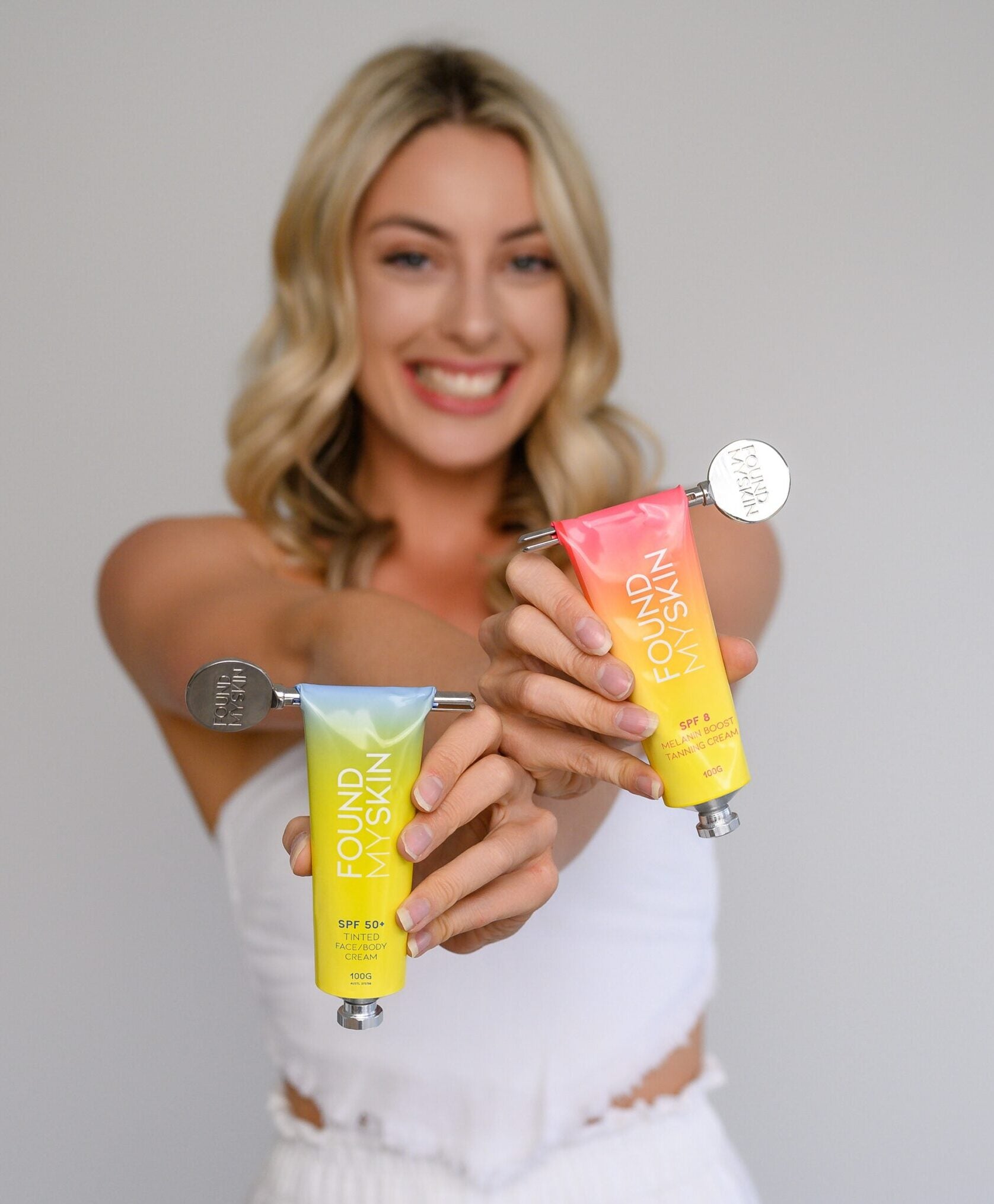 Lächelnde Frau in Weiß hält zwei FOUND MY SKIN SPF 50 getönte Tagescreme Face & Body Tuben mit Metallquetschern. Blashy Shop