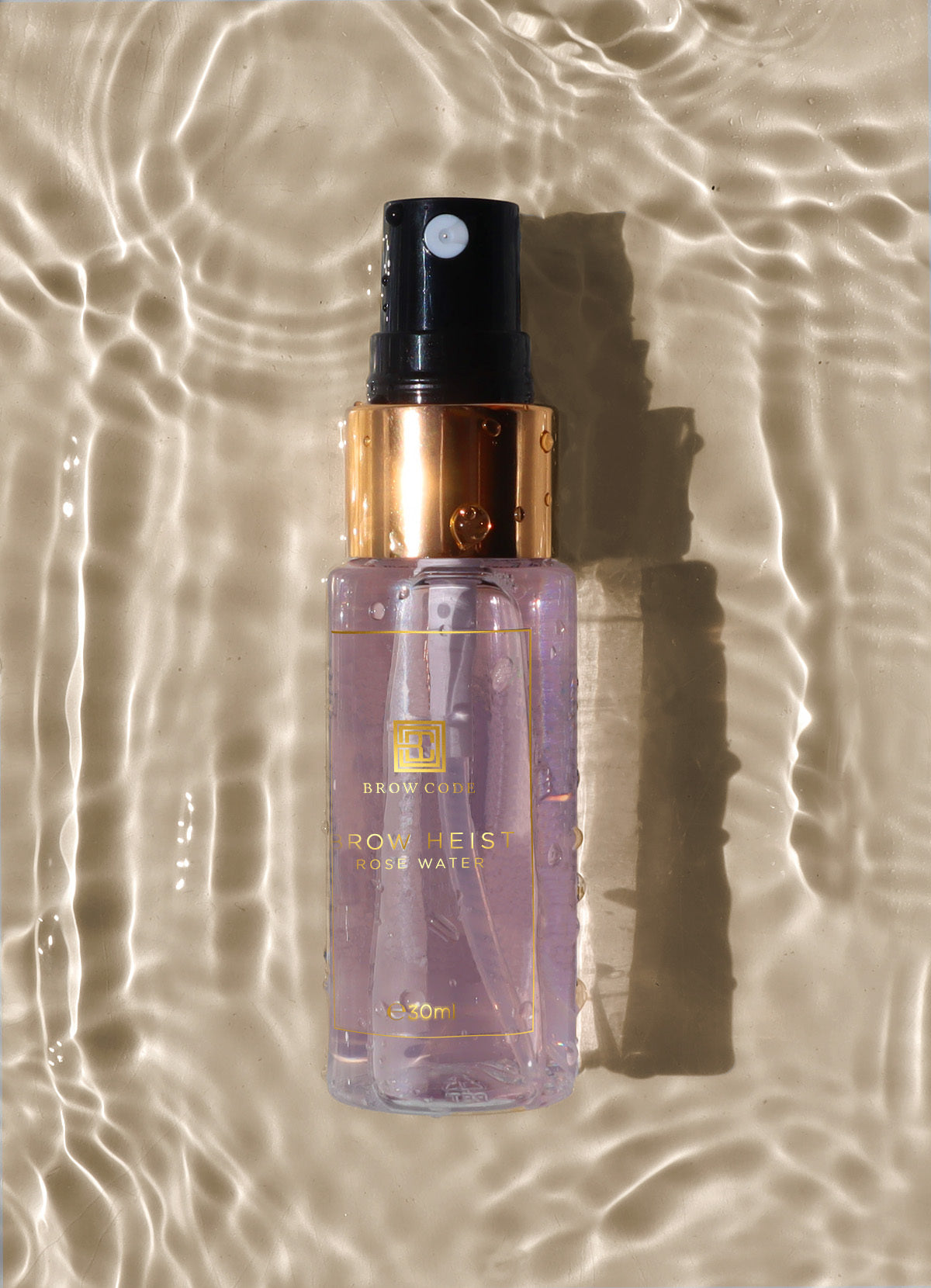 Ein Brow Code Rosenwasser Spray, 30ml, für gestylte Brauen ruht auf gewelltem Sand im Sonnenlicht. Blashy Shop