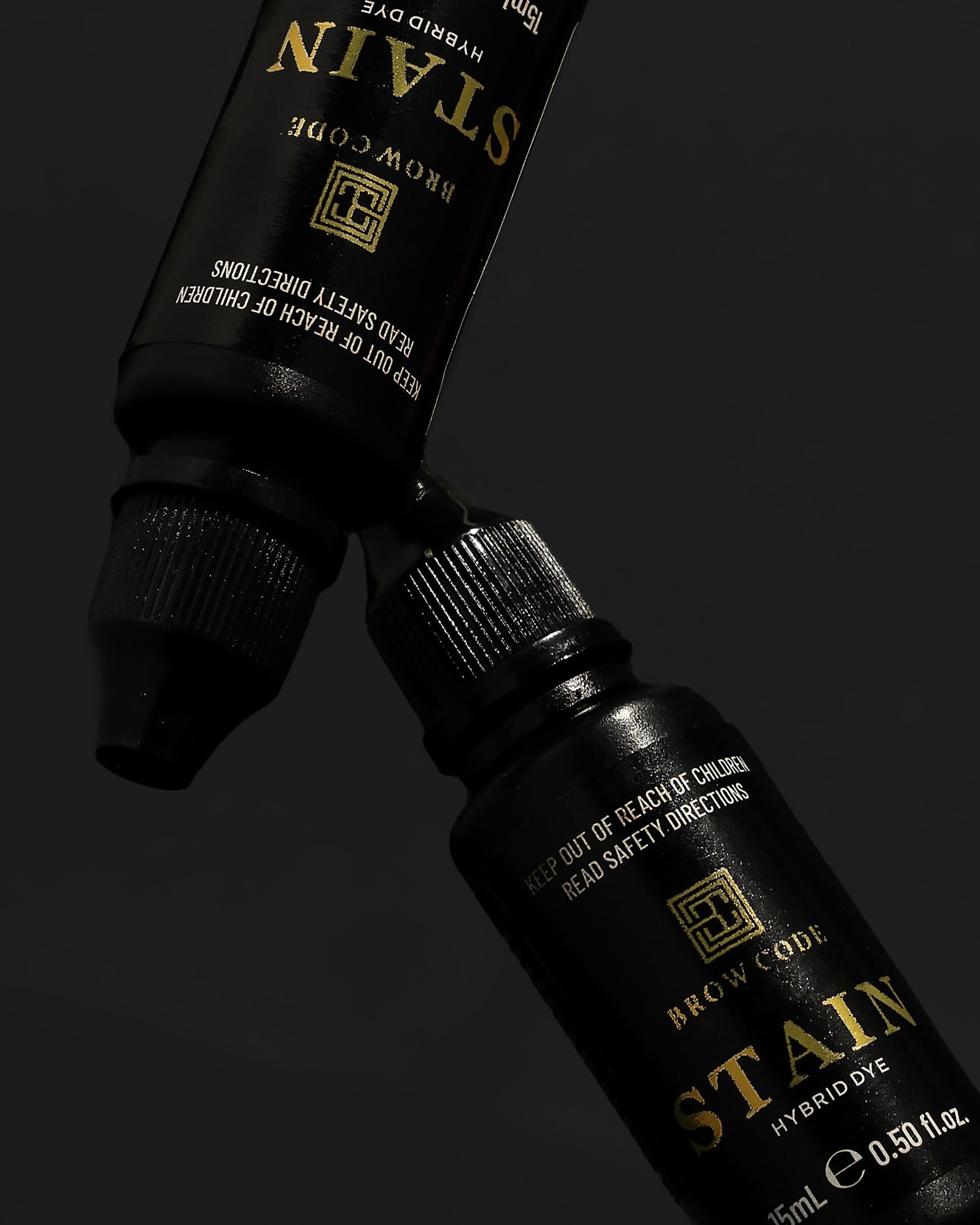 Zwei schwarze Brow Code Stain Hybrid Augenbrauenfarbe 15ml Flaschen mit goldener Beschriftung auf dunklem Hintergrund. Blashy Shop