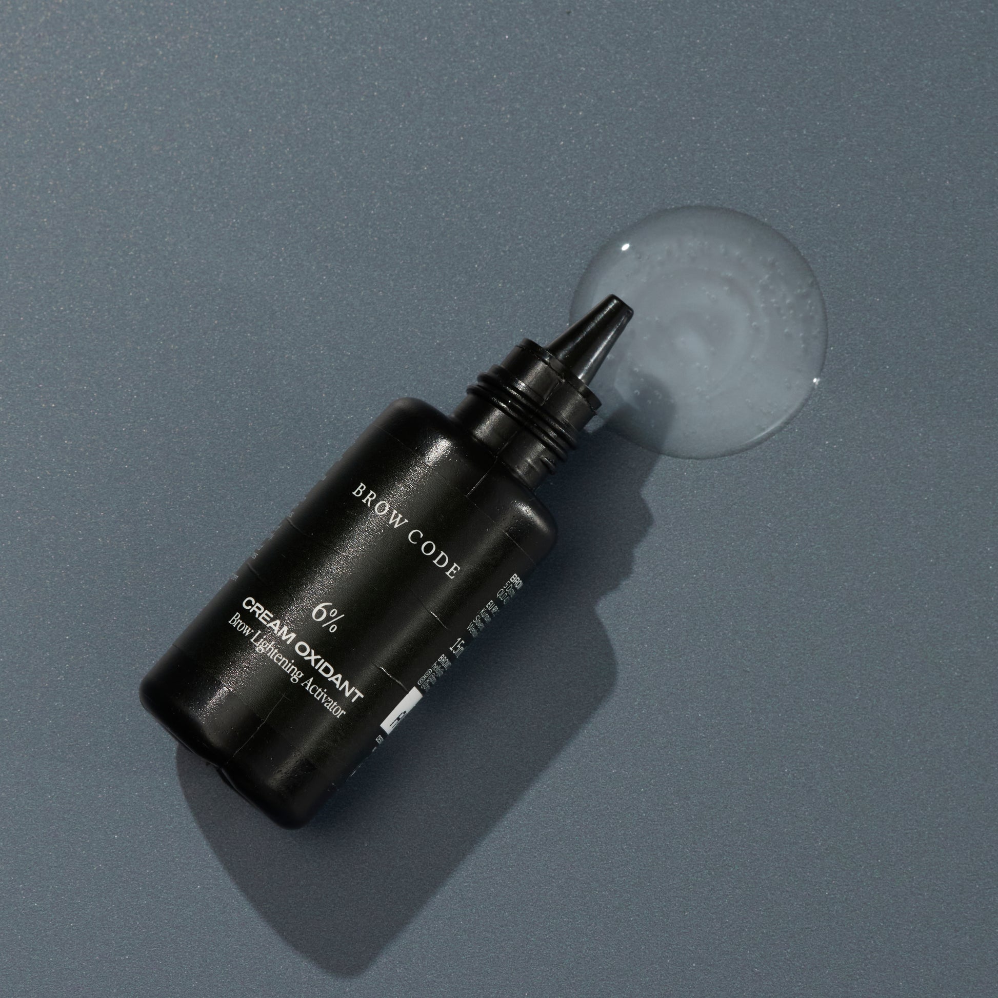 Eine schwarze Flasche Brow Code STARTER KIT Lemon Lights Augenbrauen Bleaching Kit liegt neben verschütteter Flüssigkeit auf grauem Grund. Blashy Shop