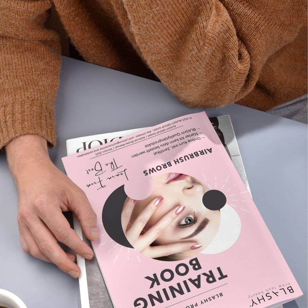 Person mit dem rosafarbenen Lash&Brow Academy Online Airbrush Brows Trainingshandbuch E-Book, das auf einem Magazin liegt. Blashy Shop