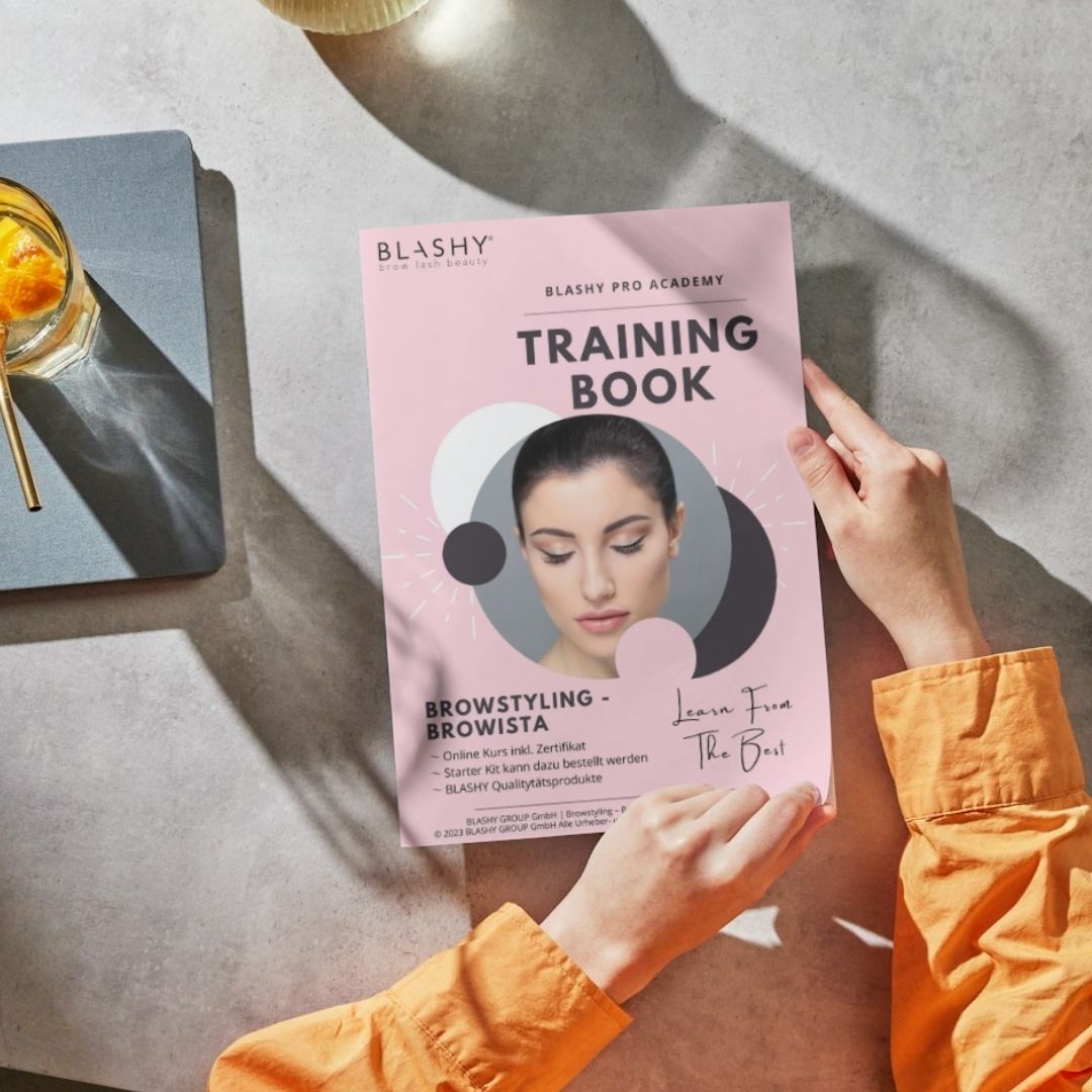 Person, die ein Trainingshandbuch - Brow Styling Browista [E-BOOK] von Lash&Brow Academy Online bei einem Orangengetränk hält. Blashy Shop