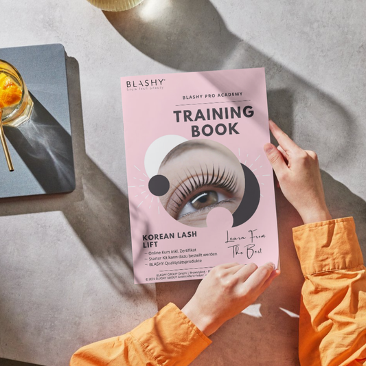 Person mit einem Trainingshandbuch - Korean Lash Lift von Lash&Brow Academy Online auf einem sonnenbeschienenen Tisch. Blashy Shop