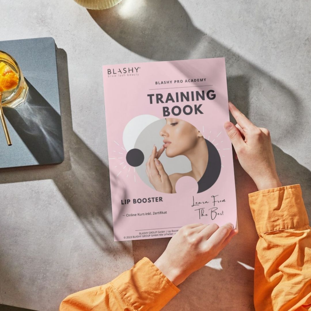 Person mit einem Lash&Brow Academy Online Lip Booster E-BOOK auf einem Tisch neben einem Getränk und einem Notizblock. Blashy Shop