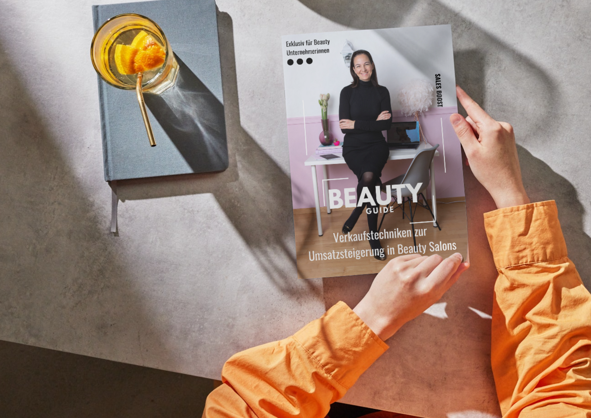 Person in orangefarbenem Hemd hält das E-Book Beauty Business Umsatzsteigerung von Lash&Brow Academy Online an einem Tisch. Blashy Shop