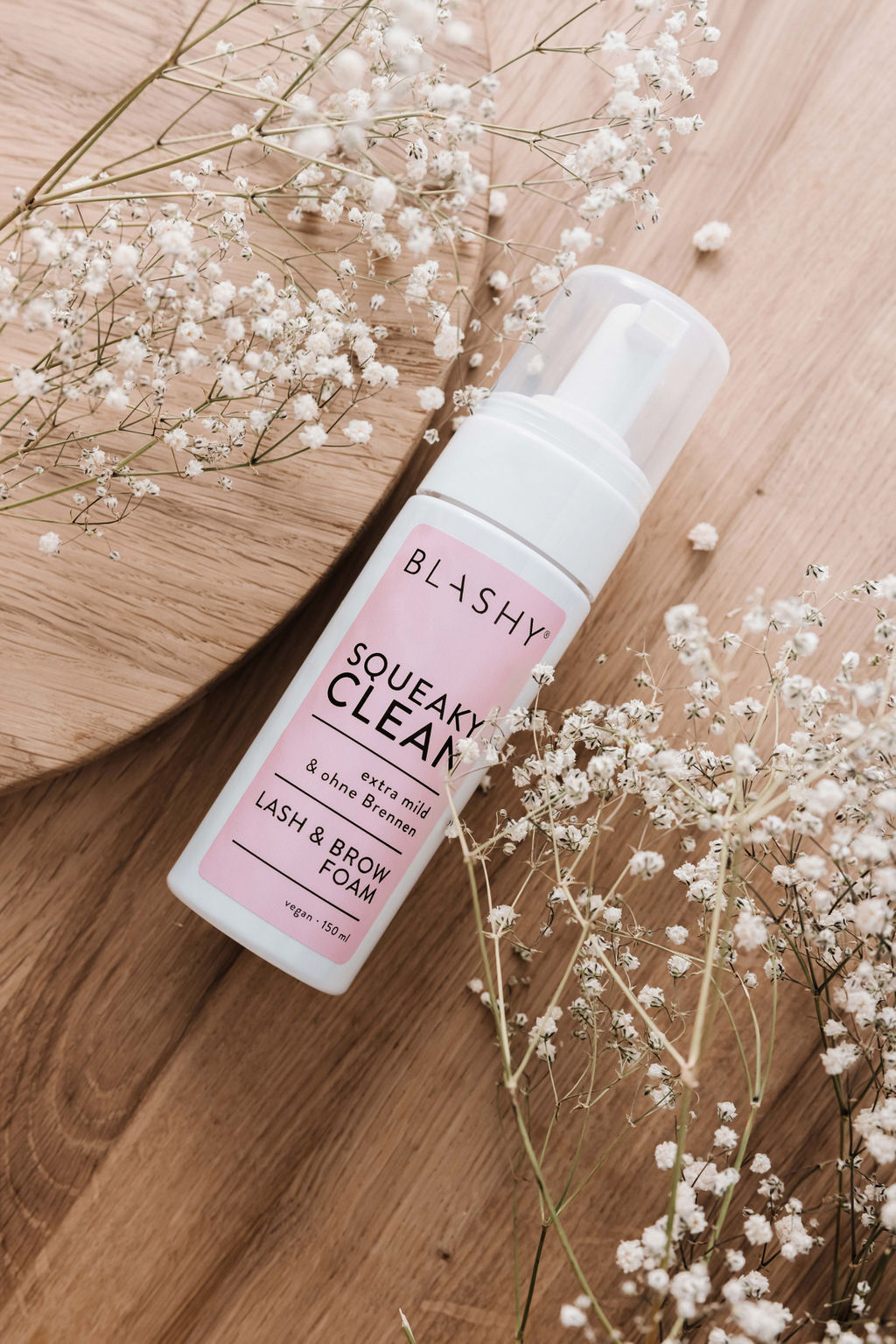 Eine Flasche Blashy SQUEAKY CLEAN Lash & Brow Schaum (60 ml) steht auf Holz, umgeben von Babys Breath Blumen. Blashy Shop